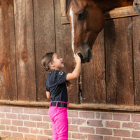A young girl in a black equestrian shirt, bright pink riding pants, and black boots gently touches the nose of a brown horse standing in a wooden stable. Hay is scattered on the ground.