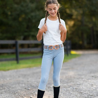 A young girl with braided hair stands on a gravel path outdoors, wearing a white t-shirt, light blue leggings with a decorative horseshoe belt, black knee-high socks, and paddock boots.