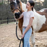 Girl in equestrian leggings seamless shirt pants standing next to a paint pony horse in equestrian riding ring
