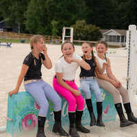 Four young equestrian girls sitting on a colorful horse pony jump in an outdoor arena wearing riding legging breeches