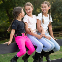 Three little girls equestrian horse back riders sitting on a dark wooden fence outdoors, wearing different equestrian-style outfits including black and bright pink boots, white shirts, and hot pink, periwinkle and light blue leggings