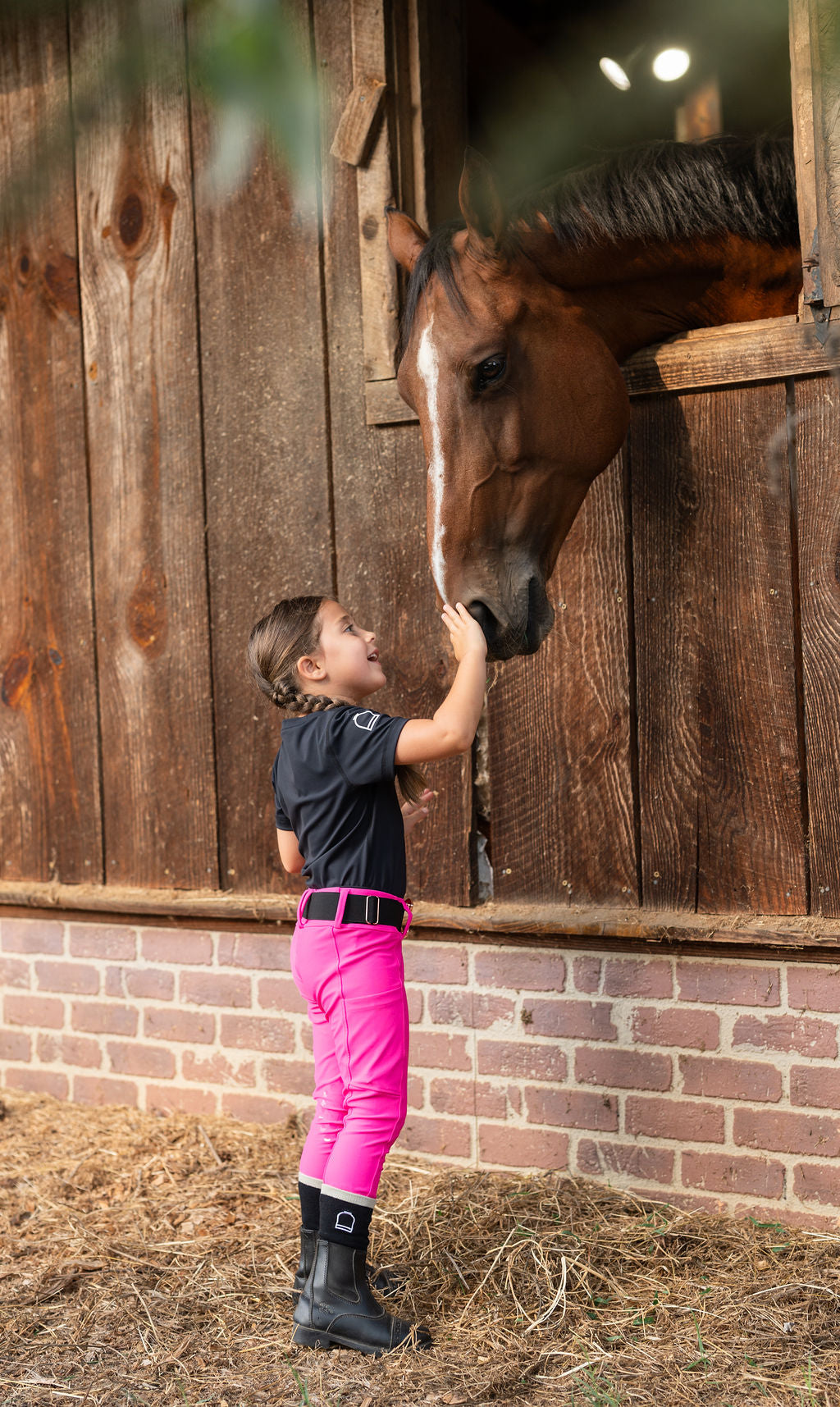 A young girl in a black equestrian shirt, bright pink riding pants, and black boots gently touches the nose of a brown horse standing in a wooden stable. Hay is scattered on the ground.