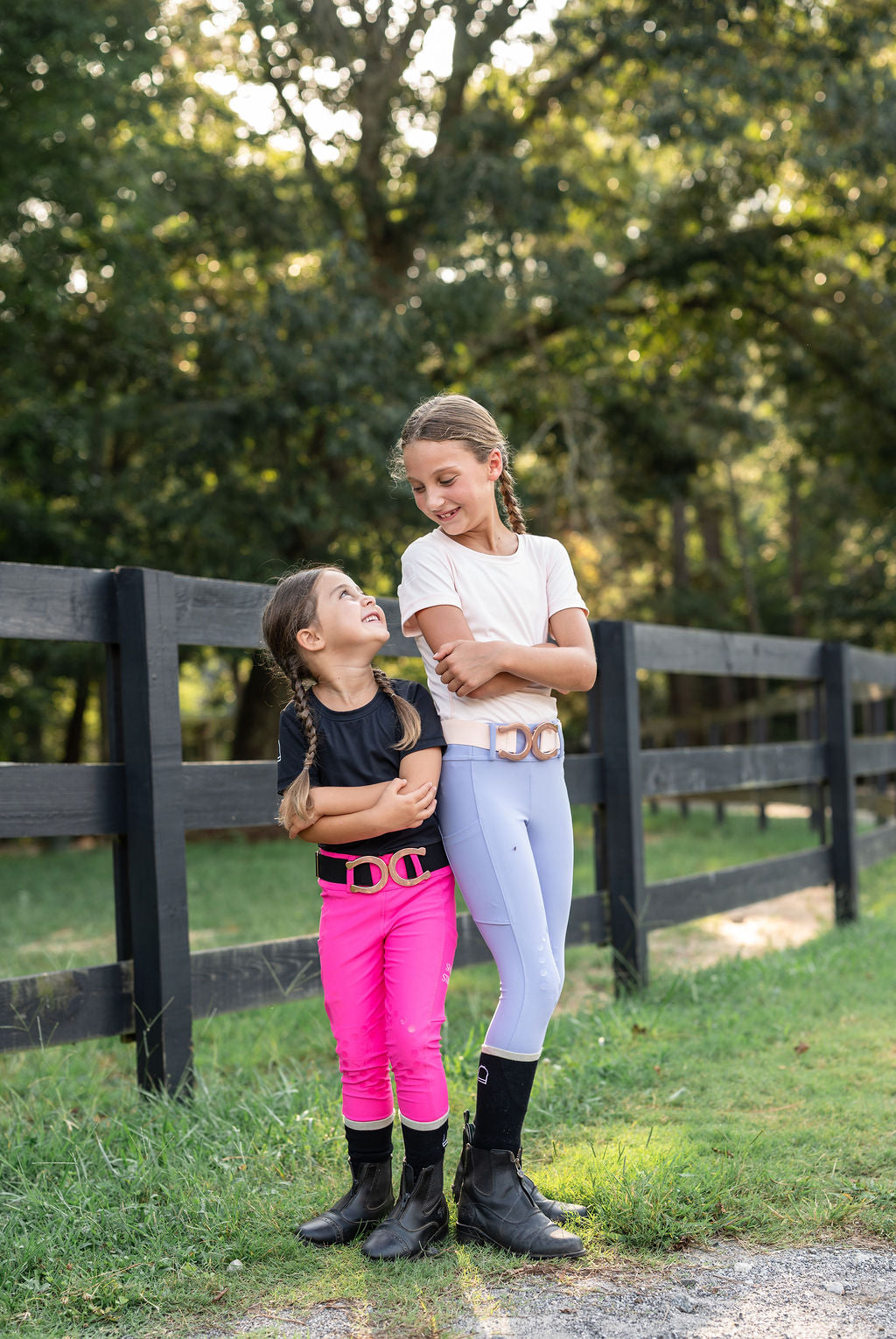 Two young girls standing outdoors in front of a black wooden fence and trees. One girl wears a white top, light blue periwinkle and hot pink cotton candy riding breeches, and black boots with a metallic belt.