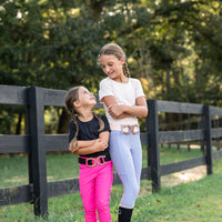 Two young girls standing outdoors in front of a black wooden fence and trees. One girl wears a white top, light blue periwinkle and hot pink cotton candy riding breeches, and black boots with a metallic belt.