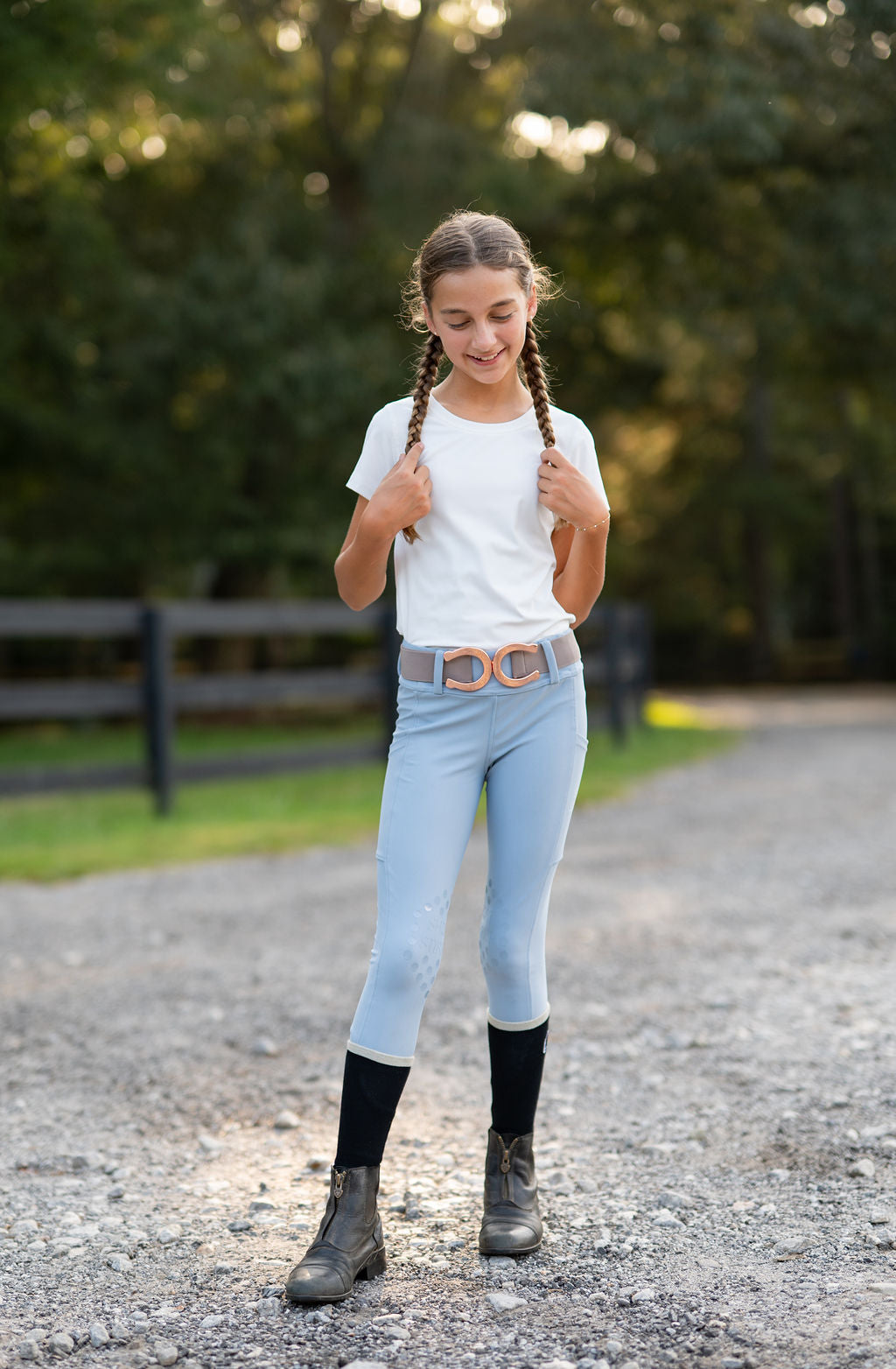 A young girl with braided hair stands on a gravel path outdoors, wearing a white t-shirt, light blue leggings with a decorative horseshoe belt, black knee-high socks, and paddock boots.