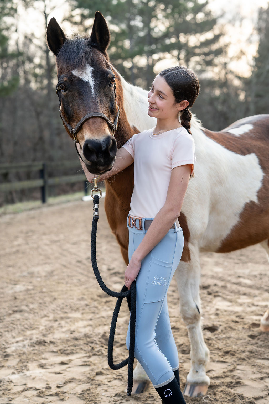 Girl in equestrian leggings seamless shirt pants standing next to a paint pony horse in equestrian riding ring