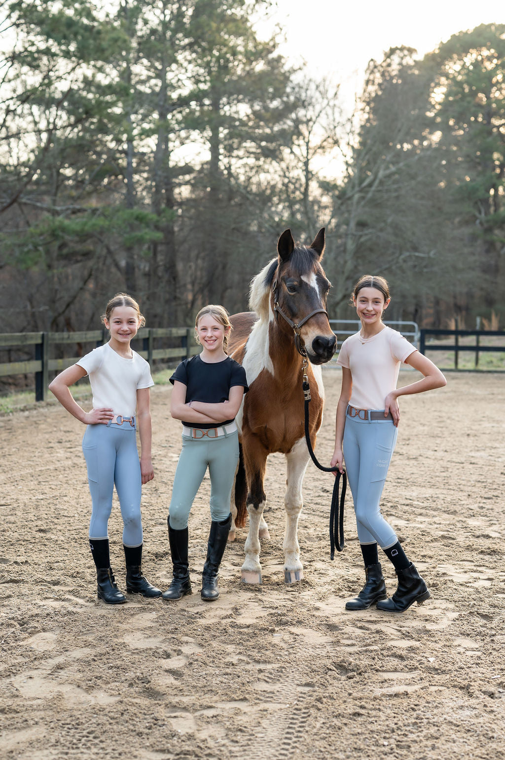 Three young girls in equestrian leggings seamless shirt pants standing next to a paint pony horse in equestrian riding ring