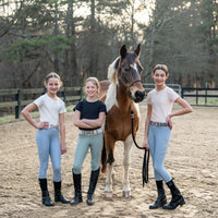 Three young girls in equestrian leggings seamless shirt pants standing next to a paint pony horse in equestrian riding ring