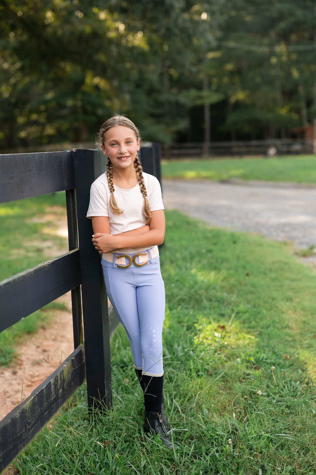 A young equestrian girl with braided hair stands outdoors leaning against a black wooden fence on a grassy field. She is wearing a white short-sleeve seamless t-shirt, light blue periwinkle riding breeches with a decorative horseshoe rose gold belt, and black boots.