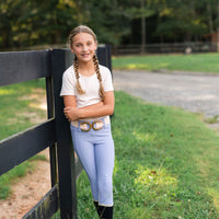 A young equestrian girl with braided hair stands outdoors leaning against a black wooden fence on a grassy field. She is wearing a white short-sleeve seamless t-shirt, light blue periwinkle riding breeches with a decorative horseshoe rose gold belt, and black boots.