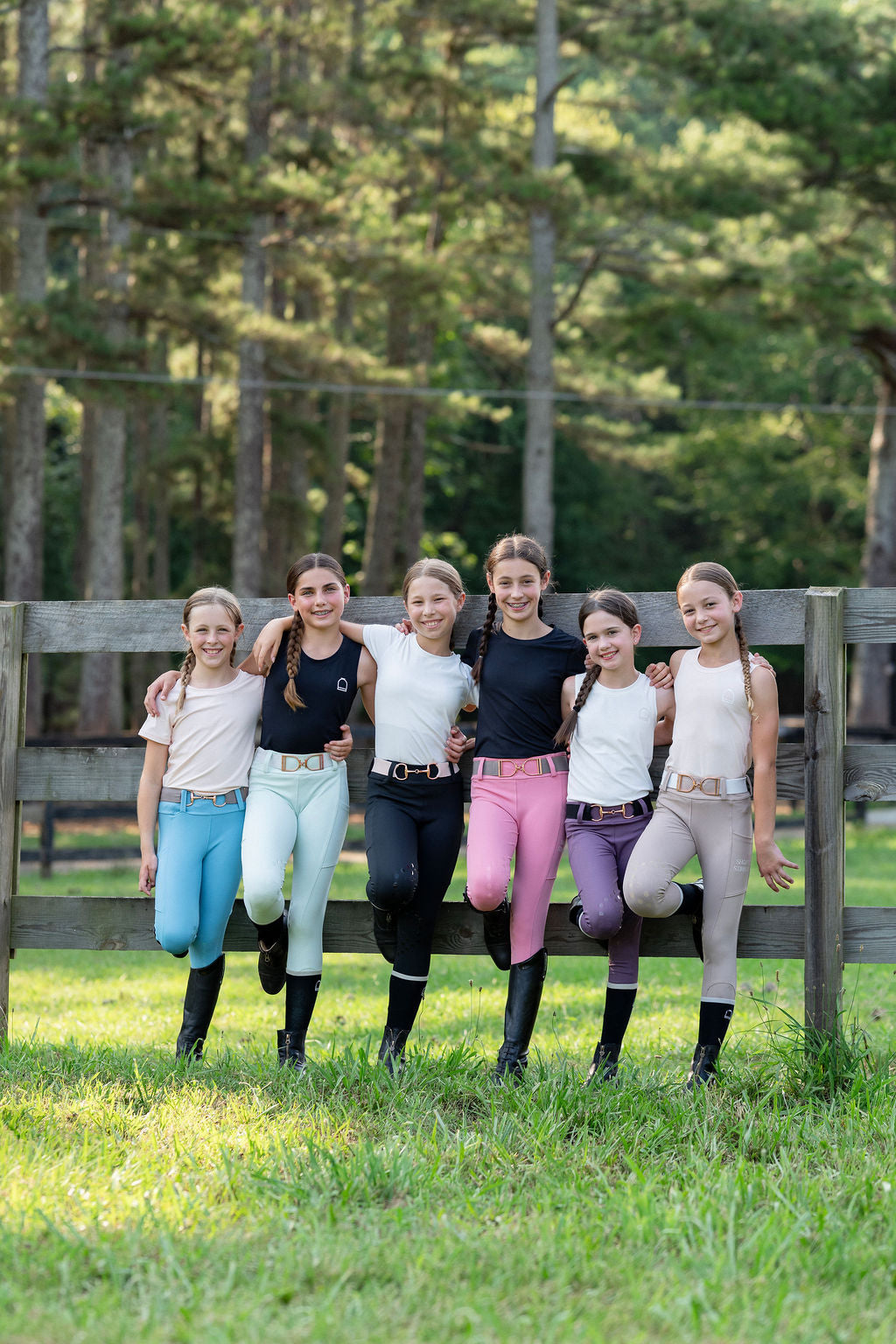 Group of five young women in equestrian attire standing in a grassy field with trees in the background.