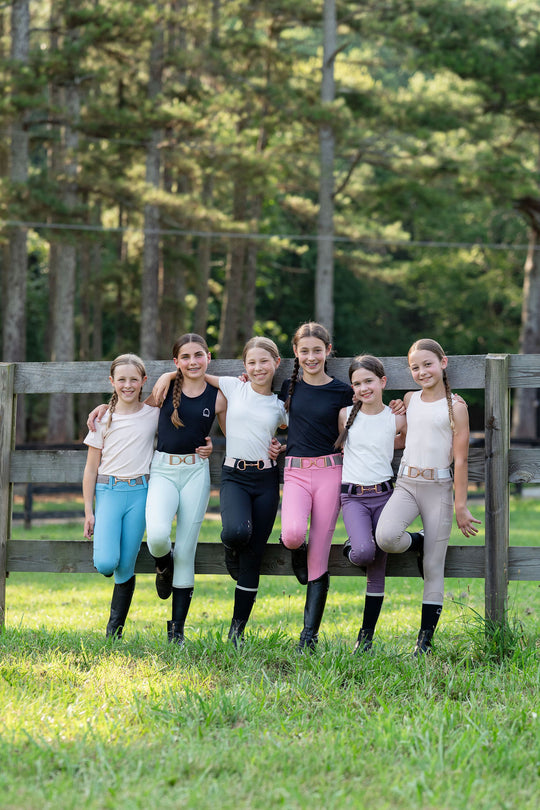 Group of five young women in equestrian attire standing in a grassy field with trees in the background.