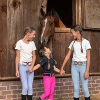 Three girls in matching equestrian outfits stand in front of a wooden stable door with a horse peeking out. The outfits feature periwinkle, hot pink and light blue riding breeches, black riding boots, and matching white black and pale pink seamless tops with decorative horseshoe rose gold belt buckles.