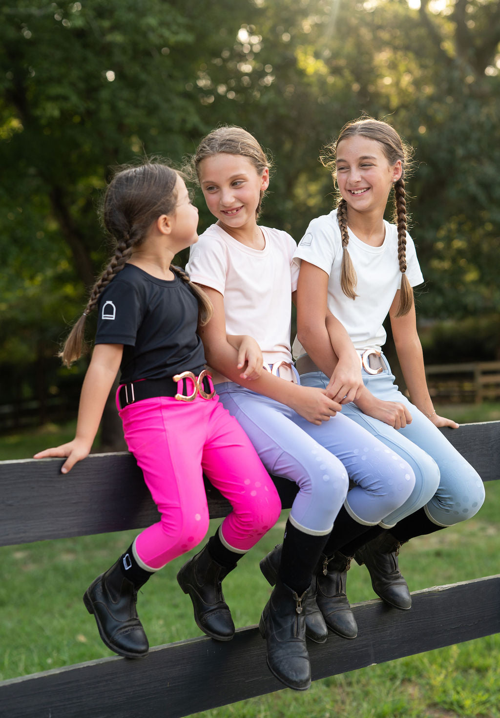Three little girls equestrian horse back riders sitting on a dark wooden fence outdoors, wearing different equestrian-style outfits including black and bright pink boots, white shirts, and hot pink, periwinkle and light blue leggings
