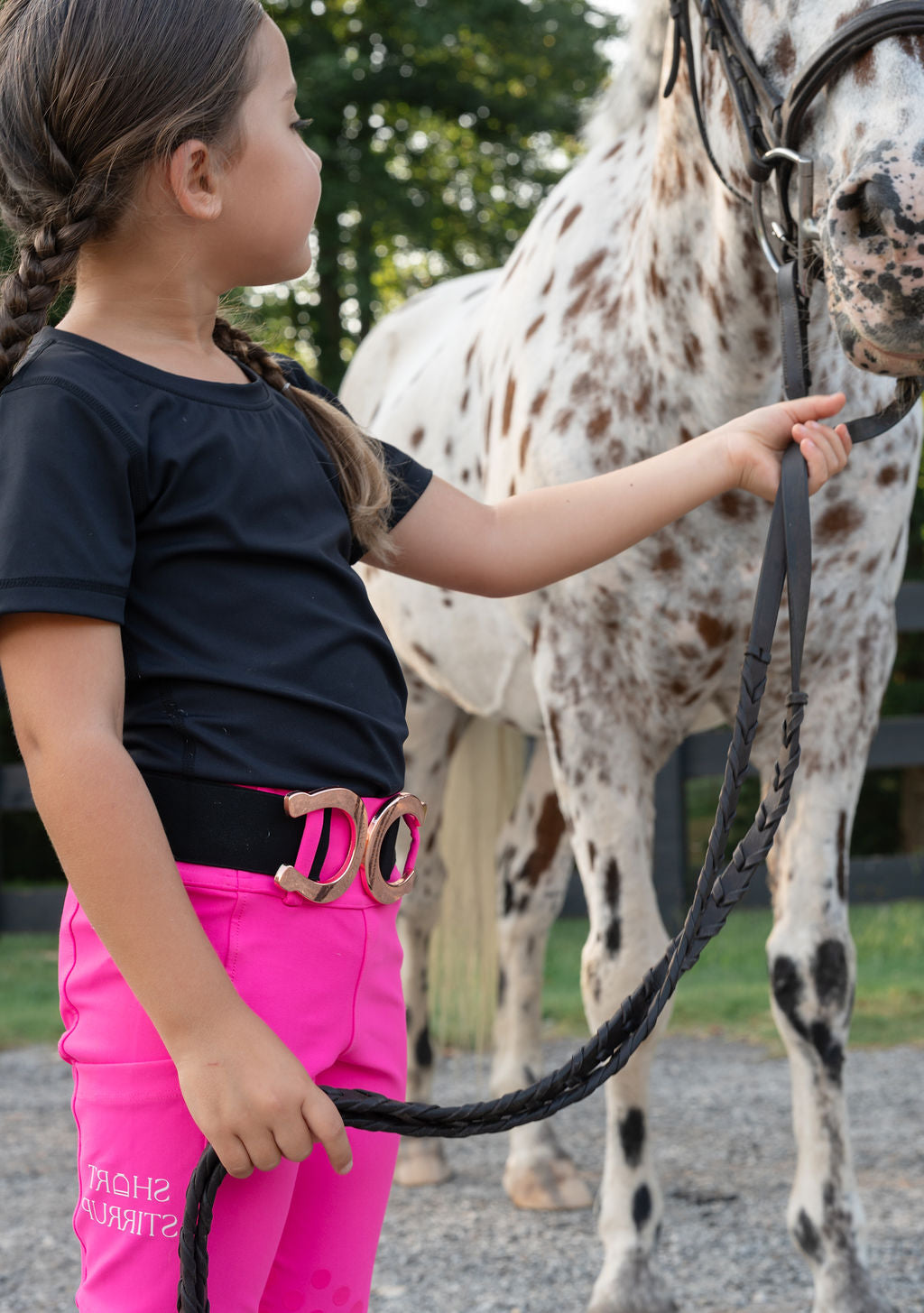 A young girl in a black t-shirt and bright hot pink riding pants with a black belt stands beside a white spotted horse, holding its reins in an outdoor paddock.