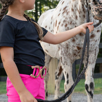 A young girl in a black t-shirt and bright hot pink riding pants with a black belt stands beside a white spotted horse, holding its reins in an outdoor paddock.