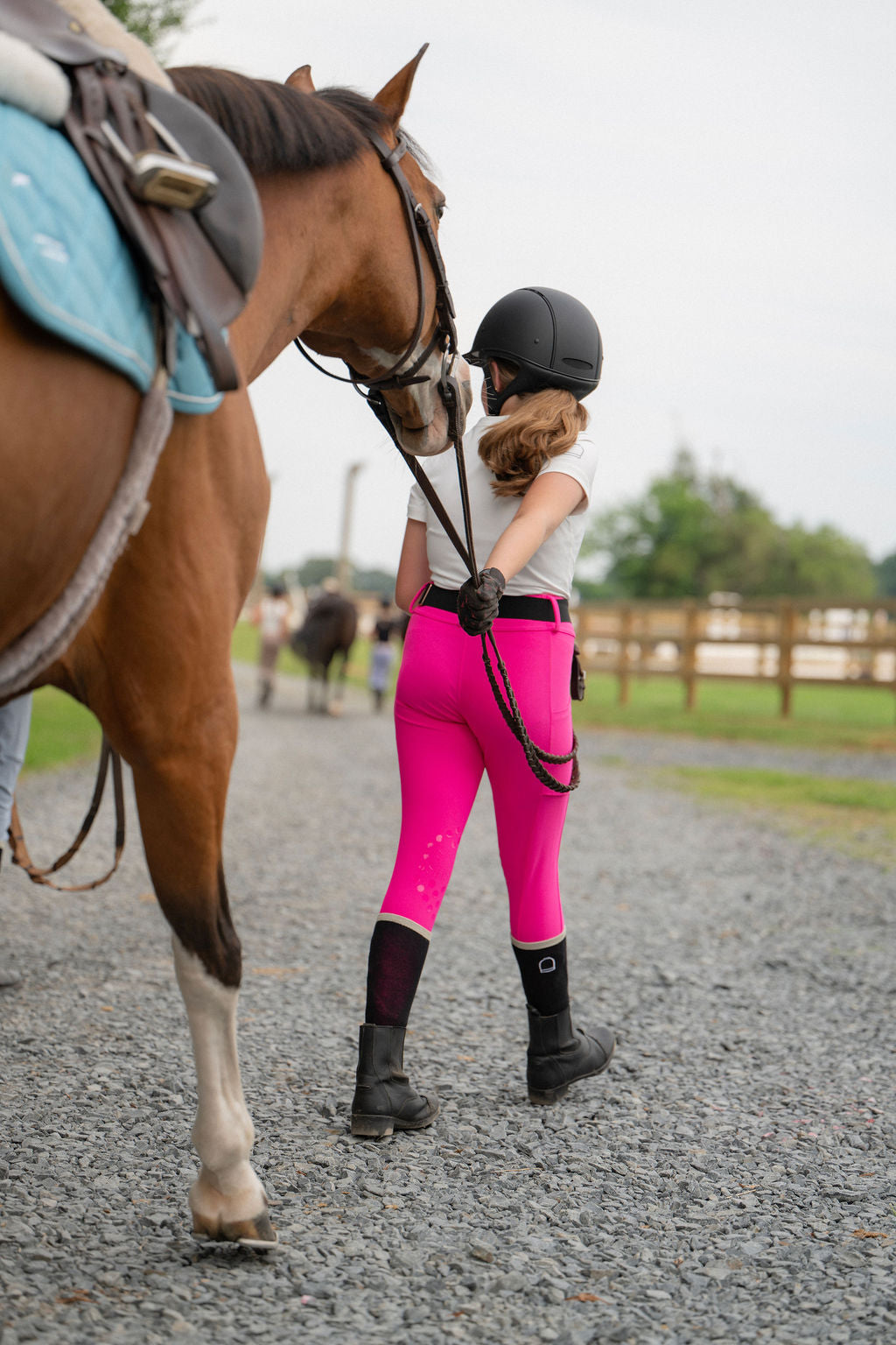 Person in pink riding legging breeches leadinng horse on a gravel path at barn